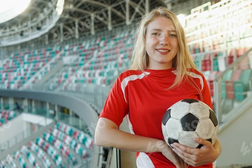 Preview: Young caucasian woman in red football strip holding ball at big soccer stadium, smiling to camera