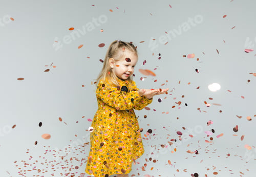 Preview: Happy little blonde girl in yellow dress catching confetti, having fun on white background