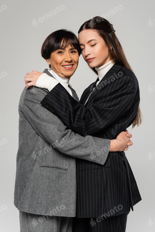 Preview: joyful asian woman embracing young and stylish daughter standing with closed eyes isolated on grey