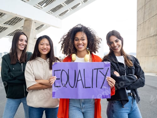 Preview: Group of cheerful multiethnic girls looking at camera holding a sign that says: Equality. Feminism,