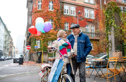 Preview: Happy senior couple with bicycle and balloons outdoors on street in city.