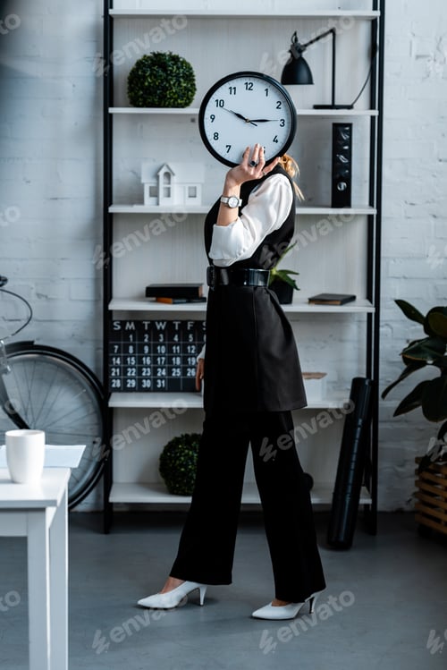 Preview: businesswoman in formal wear holding clock in front of face at workplace
