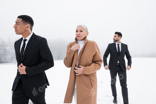 Preview: interracial bodyguards in formal wear walking near senior businesswoman on snowy field