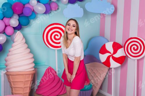 Preview: Portrait of joyful young woman in pink dress on background decorated with huge candies and ice cream