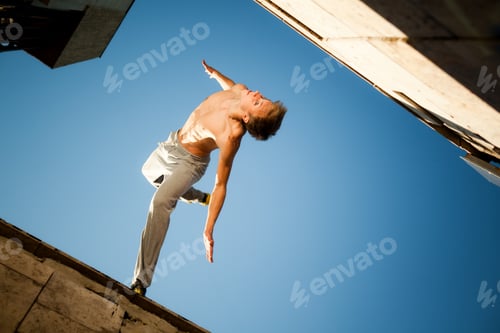 Preview: Young man jumping and practicing parkour between buildings outside on clear summer day with blue sky