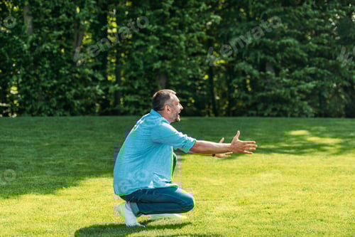 Preview: side view of happy man with open arms crouching and looking away in park