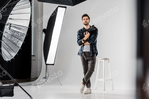 Preview: full length view of confident young photographer holding professional camera and looking at camera