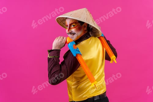 Preview: Portrait of a clown with white facial makeup on a pink background, with toy guns and a Chinese hat