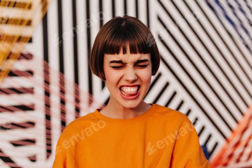 Preview: Emotional girl with brunette hair in orange sweatshirt makes funny face outside. Hipster in bright
