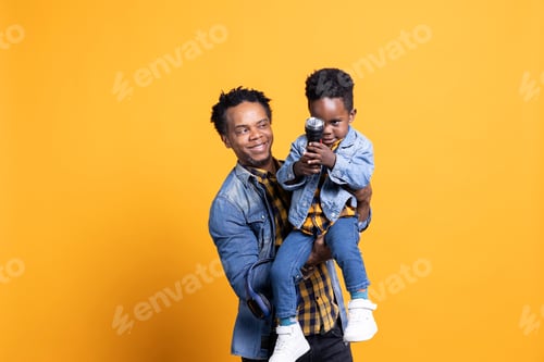 Preview: Joyful african american dad is carrying his toddler against yellow backdrop