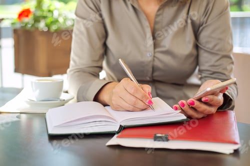 Preview: Businesswoman writing in notebook using smartphone, coffee break