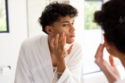 Preview: Applying skincare, teenage boy in bathrobe examining face in bathroom mirror