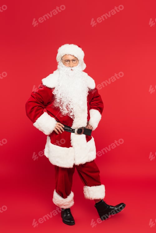 Preview: Santa claus in costume holding hands on hips on red background