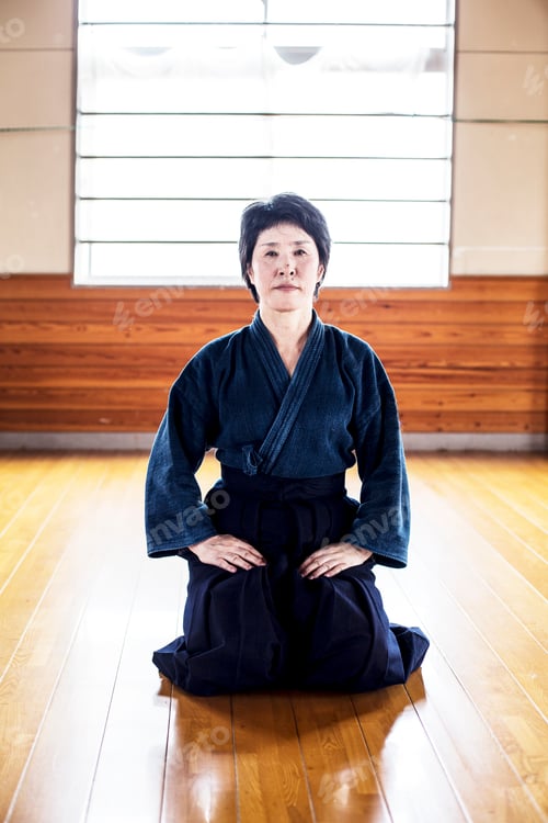 Preview: Female Japanese Kendo fighter kneeling on wooden floor, looking at camera.