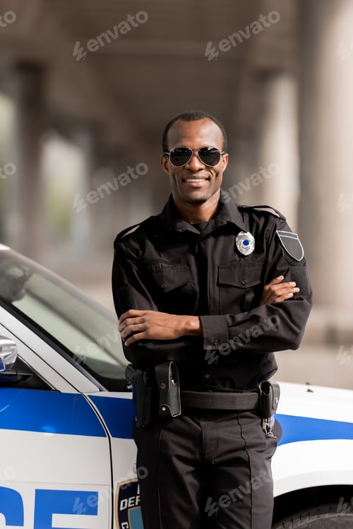 Preview: smiling african american police officer with crossed arms leaning back on car and looking at camera