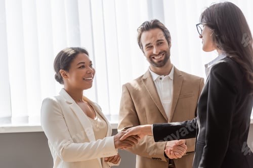 Preview: multiethnic businesswomen in formal wear shaking hands near young and smiling manager in office