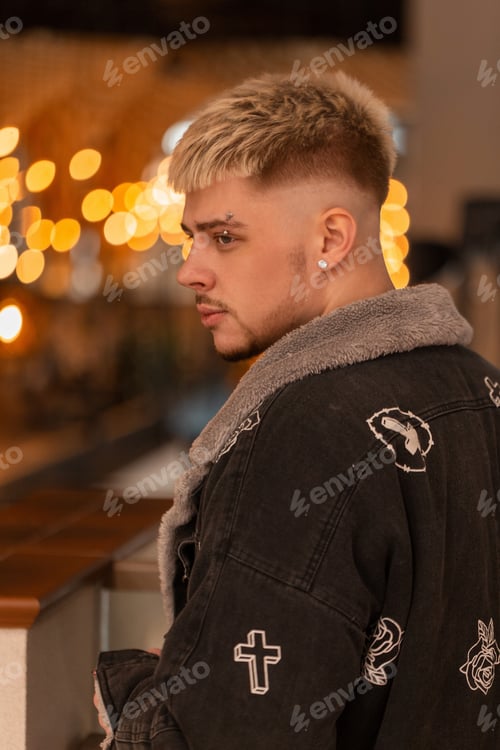 Preview: Trendy hipster guy with hairstyle in fashionable jacket walks in mall with yellow lights