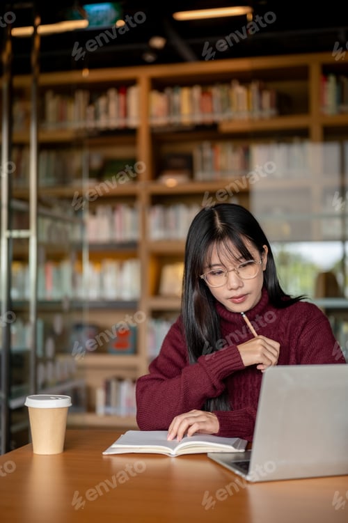 Preview: Glasses asian woman holding pencil under chin and looking at laptop aside coffee at table in library