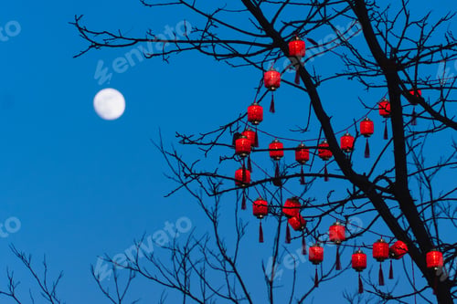 Preview: Cluster of vibrant red lanterns hanging from the branches of a tree with the moon in the night sky.