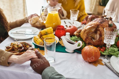 Preview: cropped multiracial man and woman holding hands and praying on Thanksgiving next to festive dinner