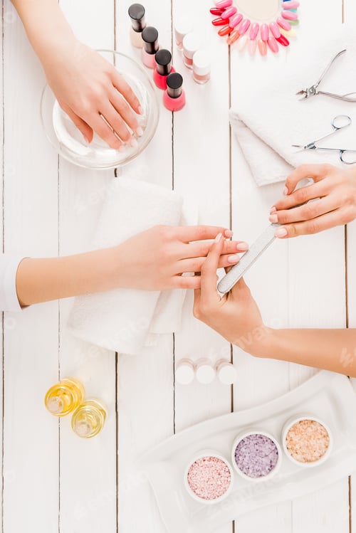 Preview: Top view of woman dipping hand in water while manicurist using nail file