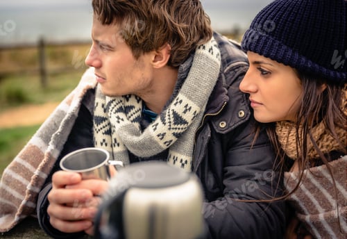 Preview: Young couple under blanket having hot drink in a cold day