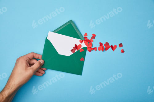 Preview: cropped view of man holding green envelope with blank white card and paper cut red hearts on blue