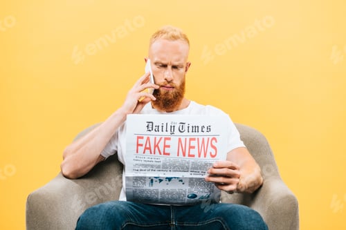 Preview: serious man reading newspaper with fake news and talking on smartphone while sitting on armchair,