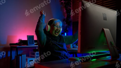 Preview: Senior woman gamer wearing headphones and playing on a desktop computer in a colorful gaming setup