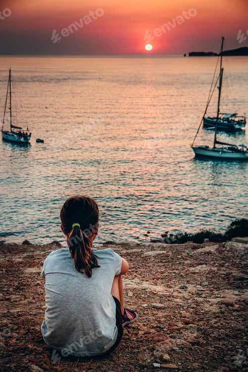 Preview: Young girl sitting by the Mediterranean sea and looking at the sunset.