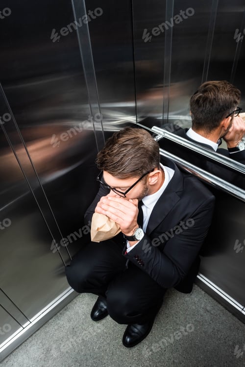 Preview: high angle view of scared businessman in suit with claustrophobia breathing in paper bag in elevator