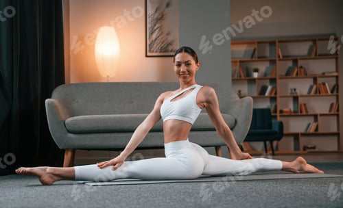 Preview: Practicing on the floor. Young woman with slim body type in white sportswear is indoors doing yoga