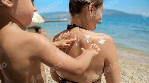 Preview: Closeup of little toddler boy rubbing sunscreen lotion in his mother's back on sunny sea beach