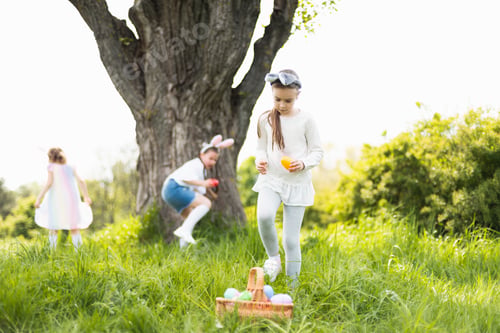 Preview: Group Of Children Wearing Bunny Ears Running To Pick Up colorful Egg On Easter Egg Hunt In Garden.