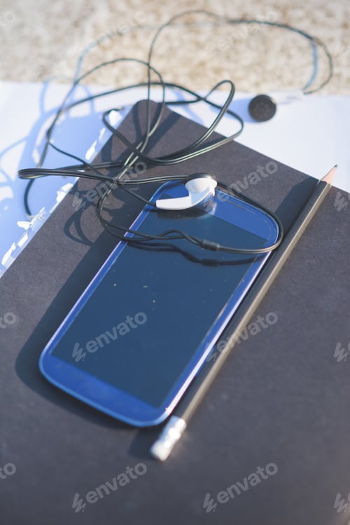 Preview: Close up on a smartphone, leaning on a sketchbook, with pencil and earphones