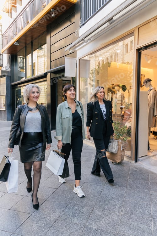 Preview: Cheerful ethnic women friends walking on pavement near shopping center with handbags in daylight