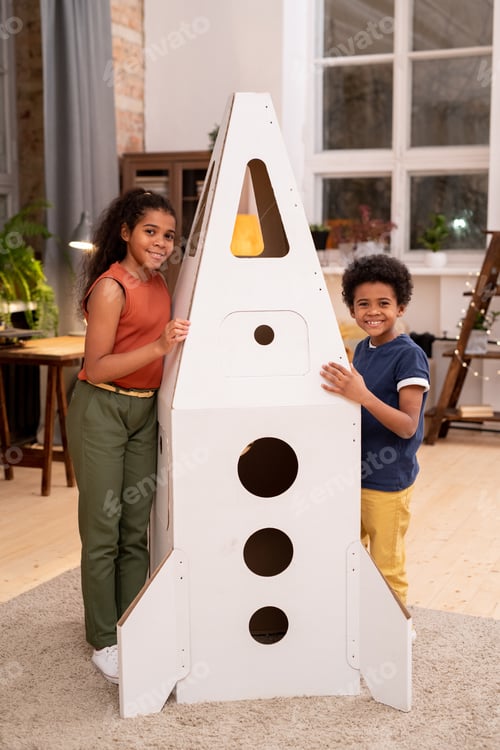 Preview: Cheerful siblings standing by big rocket and looking at camera with smiles