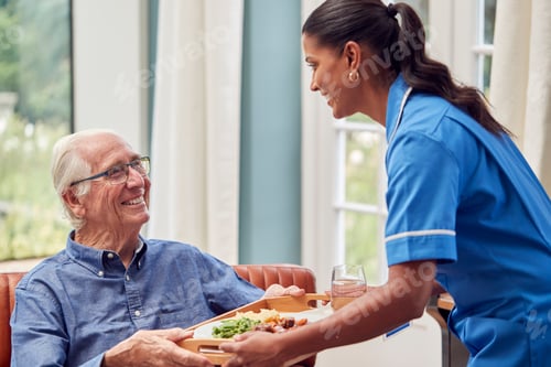 Preview: Female Care Worker In Uniform Bringing Meal On Tray To Senior Man Sitting In Lounge At Home