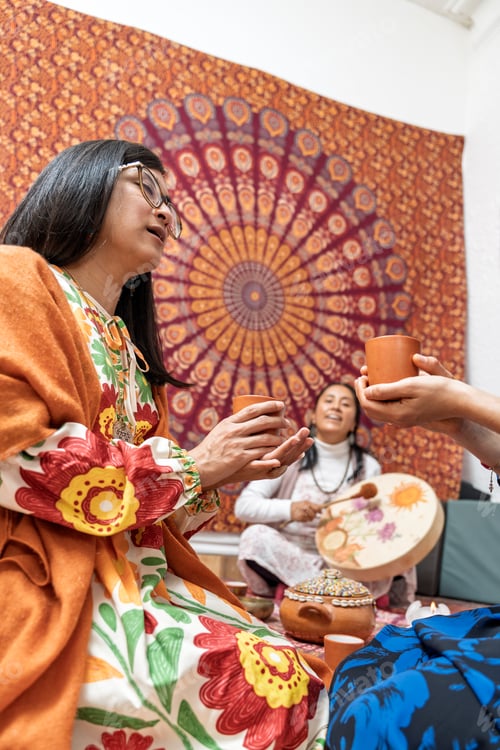 Preview: Women holding cocoa drinks during ceremony with tambourine and mandala tapestry