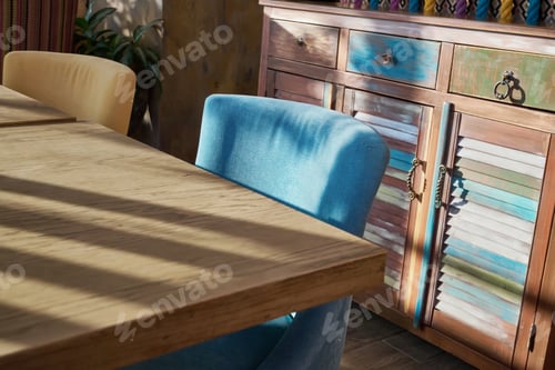 Preview: Wooden table and blue chair illuminated by sunlight next to textured colorful cabinet