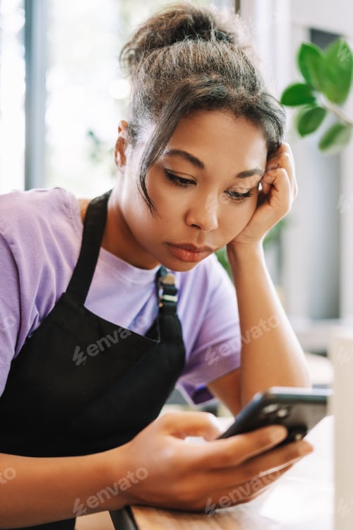 Preview: Young female coffee shop owner looking stressed while reading messages on smartphone