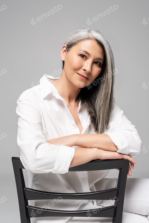 Preview: smiling asian woman with grey hair sitting on chair isolated on grey
