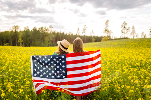 Preview: Two Young woman with american flag on blooming meadow. 4th of July. USA flag fluttering in the wind.