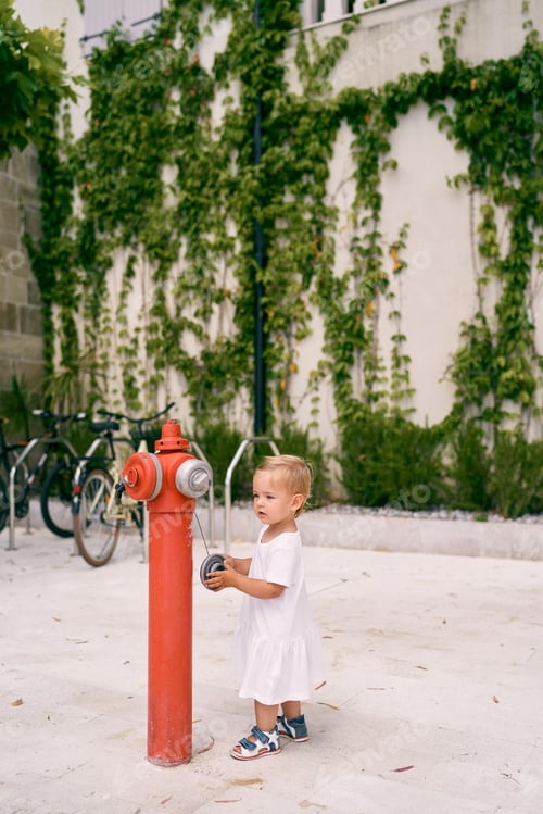 Preview: Little girl in a dress stands near a red fire hydrant in front of the building