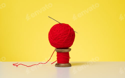 Preview: Wooden spool with red wool thread and a large needle on a white table. Handicraft items