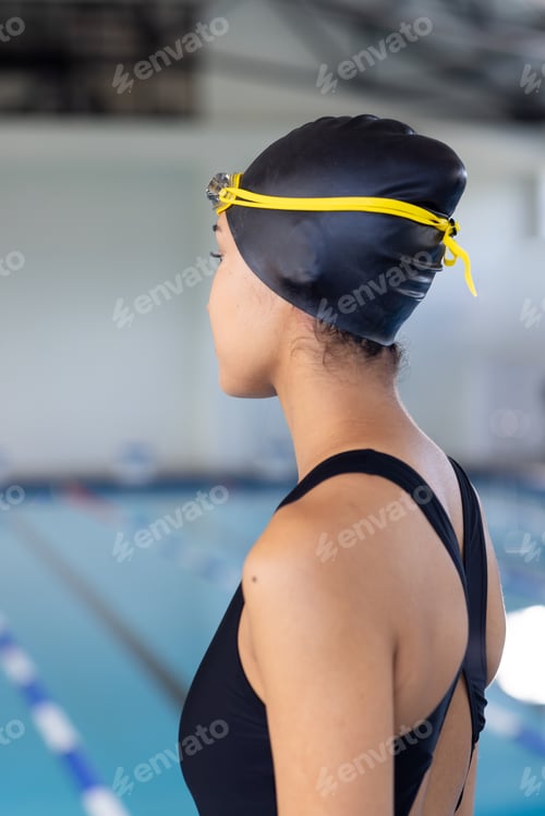 Preview: Young biracial female swimmer wearing a black swimsuit and cap, standing poolside indoors