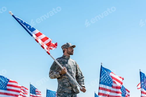 Preview: handsome soldier in military uniform and cap holding american flag while standing against blue sky