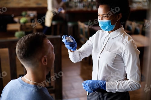 Preview: Happy waitress measuring guest's temperature with infrared thermometer in a cafe.