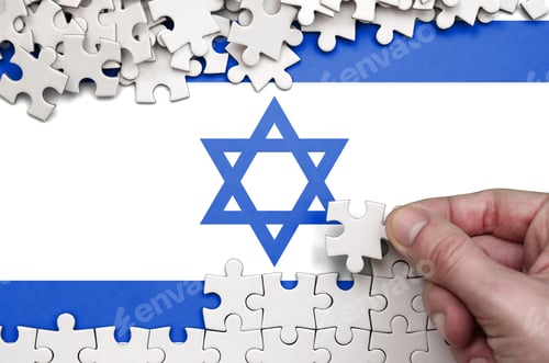 Preview: Israel flag is depicted on a table on which the human hand folds a puzzle of white color.