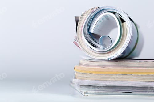 Preview: Stack of magazines with one rolled up on top, resting on a flat surface in a bright space